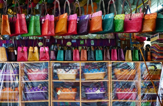 A colorful display of woven handbags arranged on shelves and hanging hooks. The bags come in vibrant shades of pink, green, orange, red, and yellow. There is a clear plastic sheet partially covering the lower shelves. The background includes a variety of additional textiles and a few payment method signs.