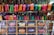 A colorful display of woven handbags arranged on shelves and hanging hooks. The bags come in vibrant shades of pink, green, orange, red, and yellow. There is a clear plastic sheet partially covering the lower shelves. The background includes a variety of additional textiles and a few payment method signs.