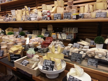 An extensive assortment of cheeses is displayed in a wooden shelving unit at a market or cheese shop. The different varieties of cheese are arranged in rows, each with labeled signs indicating their names and prices. Some cheeses are wrapped in plastic, while others exhibit a more rustic appearance. Small decorative plants can be seen among the cheeses, adding a touch of greenery to the setup.