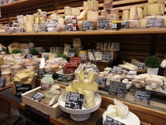 An extensive assortment of cheeses is displayed in a wooden shelving unit at a market or cheese shop. The different varieties of cheese are arranged in rows, each with labeled signs indicating their names and prices. Some cheeses are wrapped in plastic, while others exhibit a more rustic appearance. Small decorative plants can be seen among the cheeses, adding a touch of greenery to the setup.