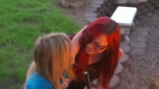Two individuals are interacting closely, one with long red hair and glasses and the other with blonde hair. They are outdoors, near a circular brick structure. The interaction appears warm and affectionate.