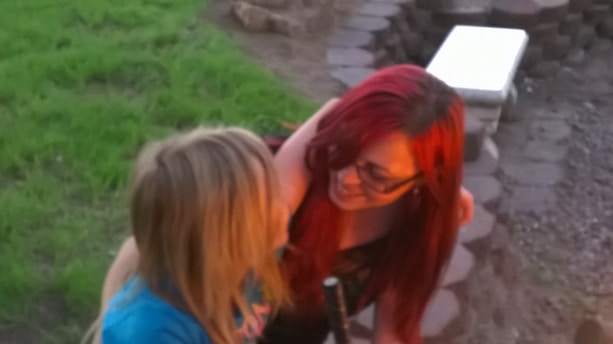 Two individuals are interacting closely, one with long red hair and glasses and the other with blonde hair. They are outdoors, near a circular brick structure. The interaction appears warm and affectionate.