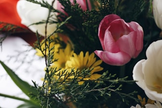 A close-up of a bouquet featuring a pink tulip, white flowers, and yellow daisies, complemented by green foliage. The composition includes a mix of delicate petals and lush greenery creating a vibrant and fresh aesthetic.
