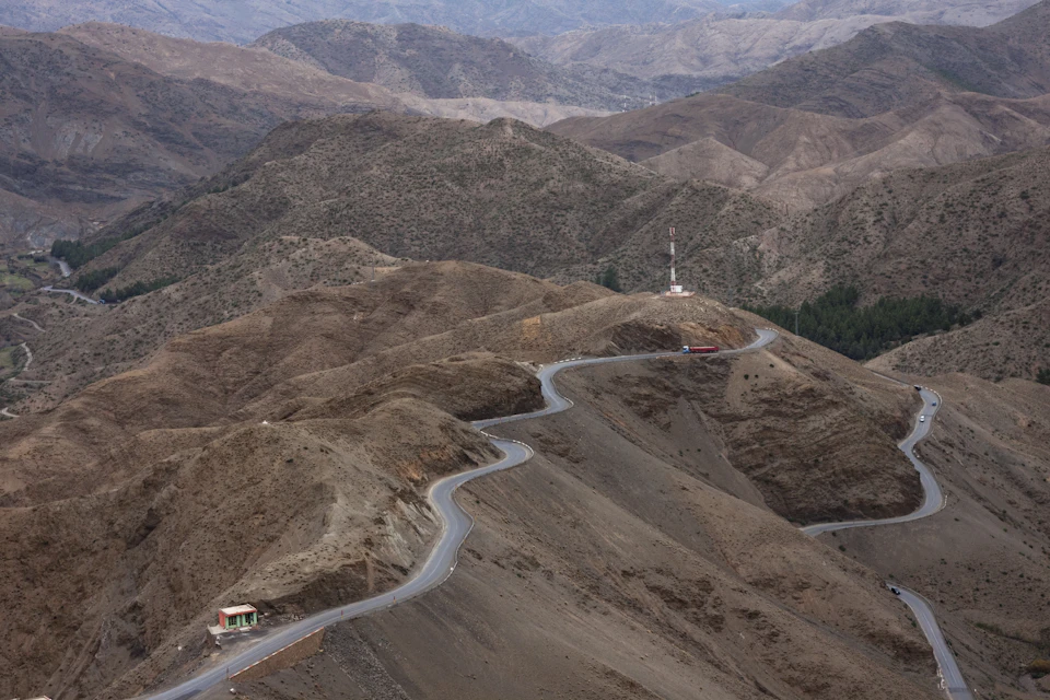 The image depicts a winding road cutting through a mountainous landscape, with two cars visible on the left side and a larger car further back on the right side. The mountainside is covered in brown dirt and features patches of green vegetation scattered throughout. In the distance, there are trees and buildings, suggesting that this location may be near a populated area or a popular tourist destination.
