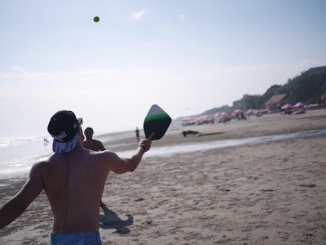 Two people are playing paddle ball on a sandy beach. One person is in the foreground, with their back to the camera, holding a paddle and facing the ocean. The sun is shining brightly, and there are beach umbrellas and people in the distance.