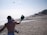 Two people are playing paddle ball on a sandy beach. One person is in the foreground, with their back to the camera, holding a paddle and facing the ocean. The sun is shining brightly, and there are beach umbrellas and people in the distance.