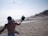 Two people are playing paddle ball on a sandy beach. One person is in the foreground, with their back to the camera, holding a paddle and facing the ocean. The sun is shining brightly, and there are beach umbrellas and people in the distance.