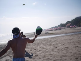 Two people are playing paddle ball on a sandy beach. One person is in the foreground, with their back to the camera, holding a paddle and facing the ocean. The sun is shining brightly, and there are beach umbrellas and people in the distance.
