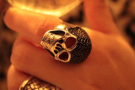 A silver skull ring with dark detailing is worn on a finger. The lighting has a warm, golden hue, accentuating the intricate design of the ring and the texture of the skin.
