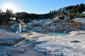 A geothermal area with steaming vents surrounded by a rocky landscape and scattered vegetation. A turquoise hot spring can be seen in the center, reflecting the light of the sun, which is low in the sky. Tall coniferous trees outline the upper ridges, adding a sense of depth and contrast to the scene.