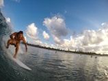 A surfer rides a wave close to the shoreline while surrounded by blue skies and fluffy white clouds. The ocean water glistens beneath him, and a few other surfers can be seen in the distance.