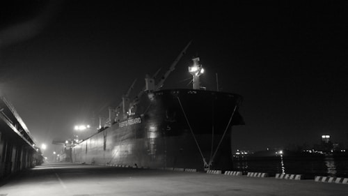 A large cargo ship is docked at a harbor during nighttime, with several cranes visible on its deck. The area is dimly lit by streetlamps, creating a stark contrast with the dark sky. Reflections from the water add a moody atmosphere, emphasizing the solitude and stillness of the scene.