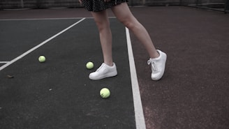 A pair of legs wearing white sneakers stands on a tennis court surrounded by several yellow tennis balls. The tennis court is divided by white lines, and the person's legs are partially visible with a black dotted skirt.