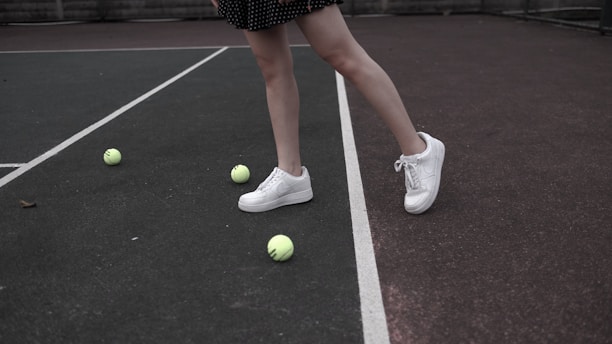 A pair of legs wearing white sneakers stands on a tennis court surrounded by several yellow tennis balls. The tennis court is divided by white lines, and the person's legs are partially visible with a black dotted skirt.