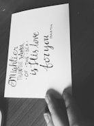 A monochromatic image featuring a handwritten quote on a piece of paper placed on a wooden surface. The quote reads 'Mightier than the waves of the sea is His love for you. Psalm 93:4'. A hand is partially visible holding the paper.