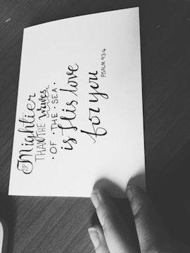 A monochromatic image featuring a handwritten quote on a piece of paper placed on a wooden surface. The quote reads 'Mightier than the waves of the sea is His love for you. Psalm 93:4'. A hand is partially visible holding the paper.