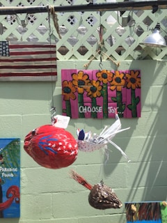 The image features an outdoor art display with various decorative elements hung against a light green wall. There is a painted wooden American flag, a vibrant artwork with sunflowers against a pink background with the words 'CHOOSE JOY', and several hanging ornaments made from natural materials, including a red object decorated with stars and stripes that resembles a seed pod or abstract bird. Another piece of art, partially visible, contains the phrases 'PARADISE FOUND'. A metal lamp hangs overhead.