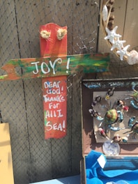 A colorful wooden cross with the words 'JOY!' and 'DEAR GOD! THANKS FOR ALL I SEA!' painted in white. It is displayed against a wire fence with various decorative seashells and starfish. Below the cross, there is a collection of painted crab figures arranged on a background that resembles newspaper. A piece of blue fabric is partially visible at the bottom.