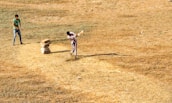 Two people are playing cricket on a dry, grassy field. One person is batting, striking the ball with a bat, while the other stands ready in the background. A makeshift wicket made of stacked stones is visible.