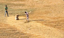 Two people are playing cricket on a dry, grassy field. One person is batting, striking the ball with a bat, while the other stands ready in the background. A makeshift wicket made of stacked stones is visible.
