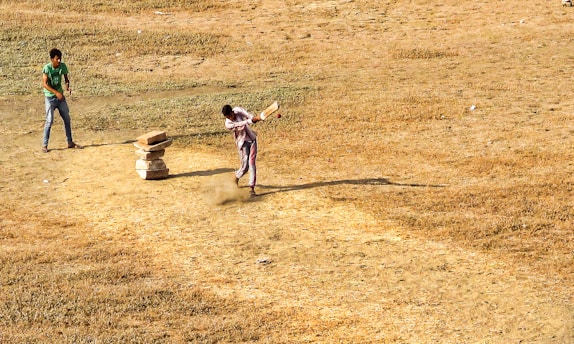 Two people are playing cricket on a dry, grassy field. One person is batting, striking the ball with a bat, while the other stands ready in the background. A makeshift wicket made of stacked stones is visible.