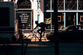 A silhouette of a cyclist riding past a shop with a prominent sign that reads 'Life is too short to wear boring shoes.' Sunlight casts a warm glow, highlighting the cyclist and parts of the storefront. A parked car and bus are visible, contributing to the urban setting.