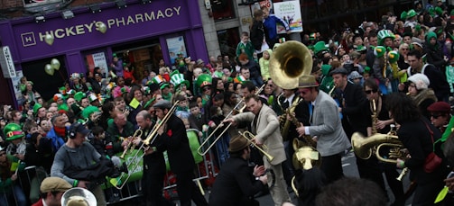 A lively street parade is taking place with numerous participants and onlookers celebrating. Many people are wearing green hats and clothes, suggesting a St. Patrick's Day celebration. A band is performing, featuring brass instruments like trombones, trumpets, and tubas. The crowd is dense and energetic, with people cheering and taking photos.