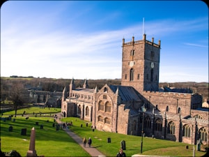 A large, historic stone cathedral with an imposing square tower featuring a clock on its fa&ccedil;ade. The cathedral is surrounded by a well-maintained grassy area with pathways, trees, and scattered gravestones. People are walking along the paths, enjoying the sunny day. The landscape extends to a distant horizon under a clear blue sky.