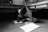 A child is sitting on the floor, deeply focused on drawing on a sheet of paper. The room is simple, with a bed and computer in the background, and shoes lined up against the wall. The image is in black and white, creating a calm and serious atmosphere.