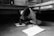 A child is sitting on the floor, deeply focused on drawing on a sheet of paper. The room is simple, with a bed and computer in the background, and shoes lined up against the wall. The image is in black and white, creating a calm and serious atmosphere.