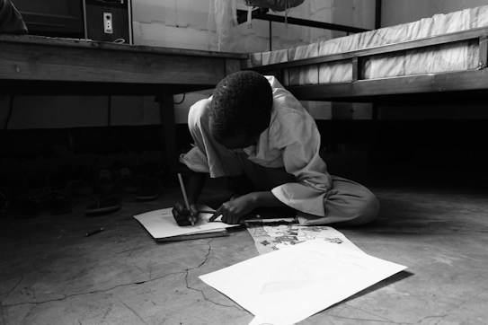 A child is sitting on the floor, deeply focused on drawing on a sheet of paper. The room is simple, with a bed and computer in the background, and shoes lined up against the wall. The image is in black and white, creating a calm and serious atmosphere.