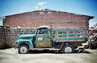 A worn-down, vintage green truck is parked in front of a stone and brick wall. The old vehicle bears signs of rust and graffiti, including various numbers and phrases. Its large wheels and metal truck bed suggest it was once used for heavy-duty purposes. Behind it, the structure is made of large stones at the bottom and red bricks at the top, with a clear blue sky above.