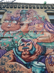 A vibrant and colorful mural covers a brick wall, featuring cartoonish characters with exaggerated features. The characters include a man with a mustache and cap, a woman with purple hair and a headband, and playful abstracts such as watermelons and a skeletal figure. The mural is heavily detailed, with various tattoo-like designs and text scattered throughout.