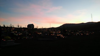 A sunset over a peaceful cemetery, with tombstones and crosses silhouetted against the colorful sky. Dimly lit candles spread across the graves create a serene atmosphere. A small building and distant hills are visible against the fading light.