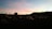 A sunset over a peaceful cemetery, with tombstones and crosses silhouetted against the colorful sky. Dimly lit candles spread across the graves create a serene atmosphere. A small building and distant hills are visible against the fading light.