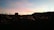 A sunset over a peaceful cemetery, with tombstones and crosses silhouetted against the colorful sky. Dimly lit candles spread across the graves create a serene atmosphere. A small building and distant hills are visible against the fading light.