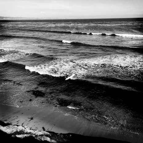 A monochrome photograph of a coastal scene capturing a lone surfer riding a wave near the shore. The ocean appears vast with gentle waves rolling towards the sandy beach. Seaweed is scattered along the shoreline, and a distant horizon is visible under an overcast sky.