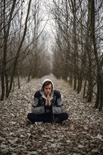 A person is sitting cross-legged in a forest of bare trees, wearing a hooded jacket. They have their hands pressed together in a meditative or prayer-like pose. The ground is covered with dry leaves, creating a serene and contemplative atmosphere.