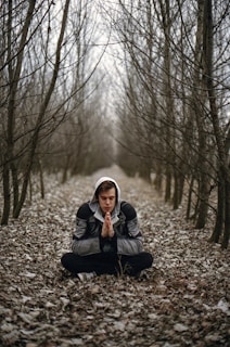 A person is sitting cross-legged in a forest of bare trees, wearing a hooded jacket. They have their hands pressed together in a meditative or prayer-like pose. The ground is covered with dry leaves, creating a serene and contemplative atmosphere.