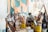 A cluttered art studio with a wooden table covered in various painting supplies, including brushes of different sizes, paint cans, bottles, and containers. The background features a partially visible abstract painting in shades of orange, teal, and white.
