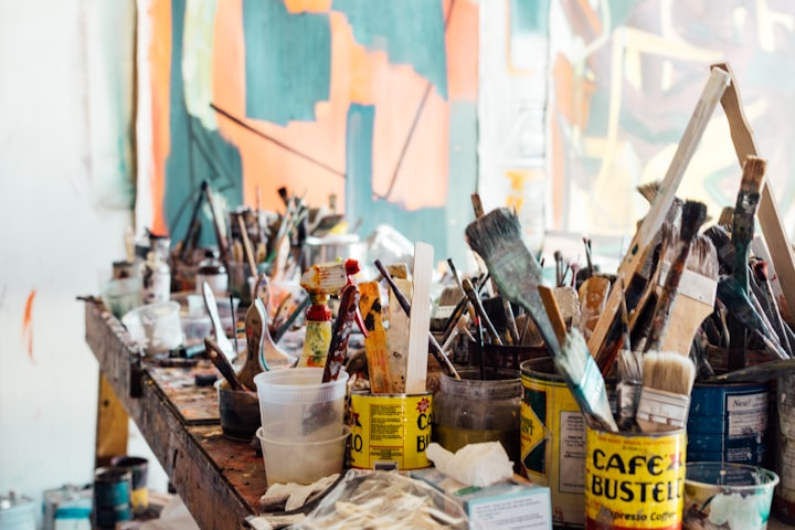 A messy artist's bench covered in paintbrushes, in front of hanging half-painted canvases.