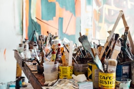 A cluttered art studio with a wooden table covered in various painting supplies, including brushes of different sizes, paint cans, bottles, and containers. The background features a partially visible abstract painting in shades of orange, teal, and white.