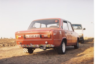A vintage classic car parked on a sunlit street with a Poor Bastard Company sticker on the bumper.