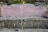 A stone bench with a pinkish concrete backrest bearing four names, separated by red lines. The bench is surrounded by natural elements like rocks, moss, and grassy vegetation.