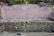 A stone bench with a pinkish concrete backrest bearing four names, separated by red lines. The bench is surrounded by natural elements like rocks, moss, and grassy vegetation.