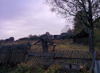 A rustic wooden fence in disrepair stretches across a grassy field, with a few broken sections. Behind the fence, there is a weathered wooden building partially obscured by trees, suggesting an abandoned or rural setting. The field and building are surrounded by overgrown vegetation, with a tree to the right adding a touch of natural beauty. The sky is overcast, casting a subdued light over the scene.