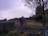 A rustic wooden fence in disrepair stretches across a grassy field, with a few broken sections. Behind the fence, there is a weathered wooden building partially obscured by trees, suggesting an abandoned or rural setting. The field and building are surrounded by overgrown vegetation, with a tree to the right adding a touch of natural beauty. The sky is overcast, casting a subdued light over the scene.