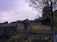 A rustic wooden fence in disrepair stretches across a grassy field, with a few broken sections. Behind the fence, there is a weathered wooden building partially obscured by trees, suggesting an abandoned or rural setting. The field and building are surrounded by overgrown vegetation, with a tree to the right adding a touch of natural beauty. The sky is overcast, casting a subdued light over the scene.