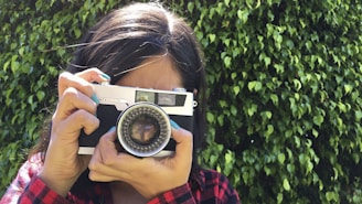 Close-up of a smiling person holding a vintage camera surrounded by plants.