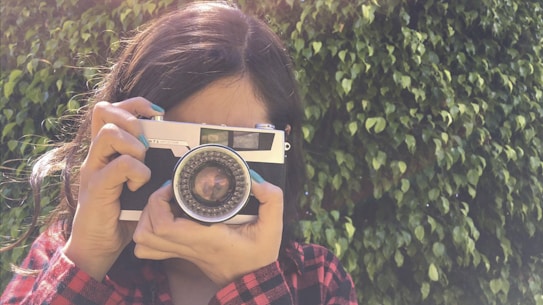 A person holding a vintage camera up to their face, capturing a photo. The background consists of lush green leaves, creating a natural setting. The individual is wearing a red plaid shirt, and their hair cascades down naturally.